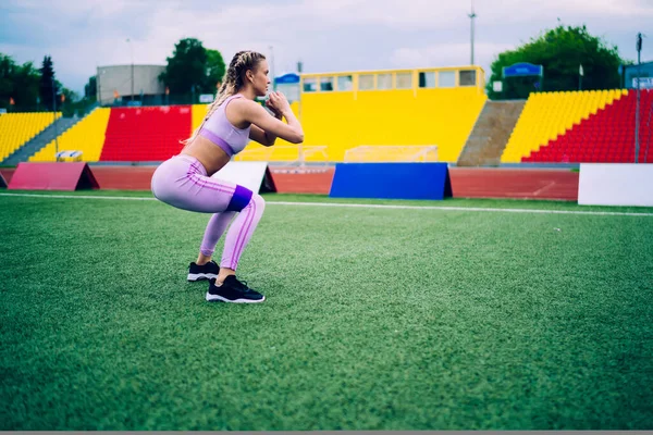 現代のスポーツにおける大人の運動女性のサイドビュー離れて見て市のスタジアムのグランドスタンドに対して緑の草の上にスクワット弾性バンドとトレーニングを持っている — ストック写真