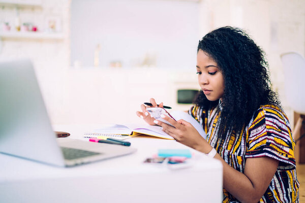 Серьезный черный подросток с вьющимися волосами просматривает социальные сети на смартфоне, сидя за столом и делая домашнее задание в уютной комнате
