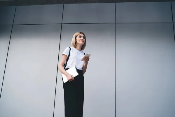 オフィスビルの近くに立って離れて見ている間 スマートフォンを使用して手にタブレットとカジュアル服の幸せなブロンドの女性 — ストック写真