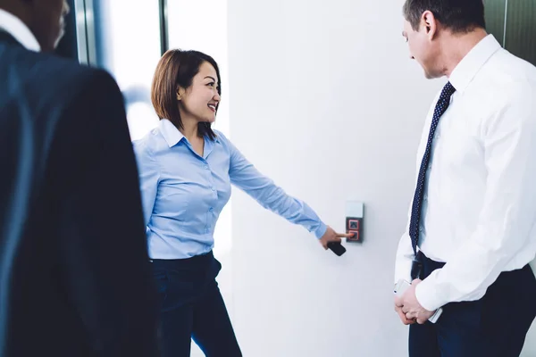 Side view of smiled Asian female in office style clothing with male colleagues having easy conversation and waiting lift while ringing call button