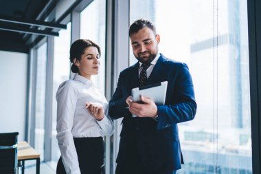 Takım elbiseli erkek yönetici, modern ofis odasının penceresinin yanında dururken tabletle kadın sekretere resmi giysiler içinde bilgi veriyor.