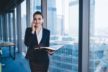 Kadın girişimci, modern gökdelenin penceresinin yanında durup kameraya bakarken iş ortağıyla cep telefonuyla konuşuyor.