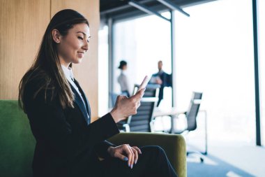 Resmi giyinen mutlu etnik bayanın yan görünüşü gülümseyen ve ofisin bulanık arka planında dinlenirken modern akıllı telefonu kullanan