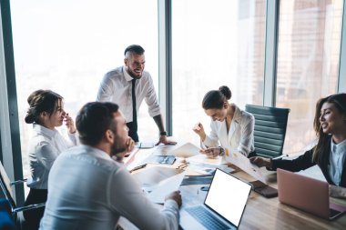 Takım lideri ayakta durup gülerek çalışma sürecinden bahsederken meslektaşları masada oturuyor ve ışık ofisinde notlar alıyor. 