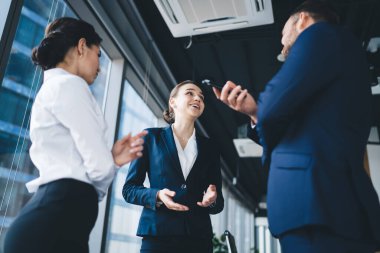 Şık takım elbiseli mutlu bir kadının modern ofiste ayakta dikilirken erkek ve kadın iş arkadaşlarıyla el kol hareketleri yapması ve konuşması.