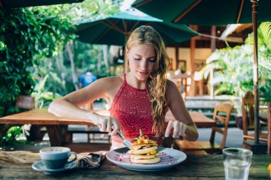 Kırmızı tişörtlü yetişkin bir kadın meyve ve meyveli lezzetli krepler yerken açık havada Bali tatil köyündeki büyük turkuaz şemsiyenin altında kafenin terasında bir fincan sıcak içecekle ahşap masada oturuyor.