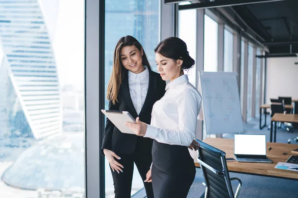 Side view of cheerful elegant businesswomen in formal wear standing in office browsing tablet in corporation against desk with blank laptop screen