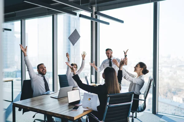 Group of excited business people with raised hands celebrating successful project tossing up papers using laptops and smartphone in modern office