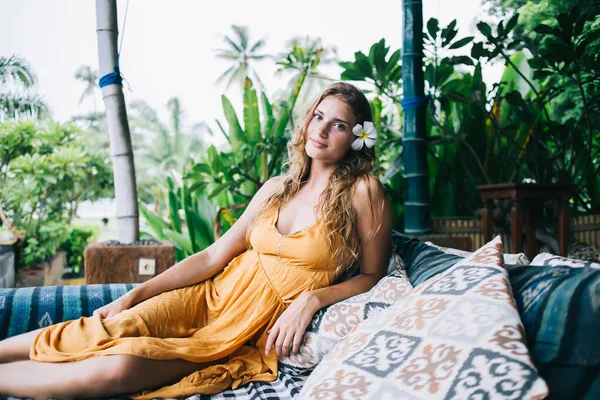 Pleased relaxed adult blond female in orange clothes with orchid in hair resting among many cushions on terrace against tropical garden at Bali resort during sunny day