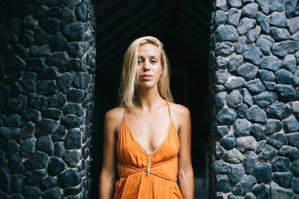Serious young blonde long haired woman wearing orange dress with straps and deep neckline standing between dark grey stone walls