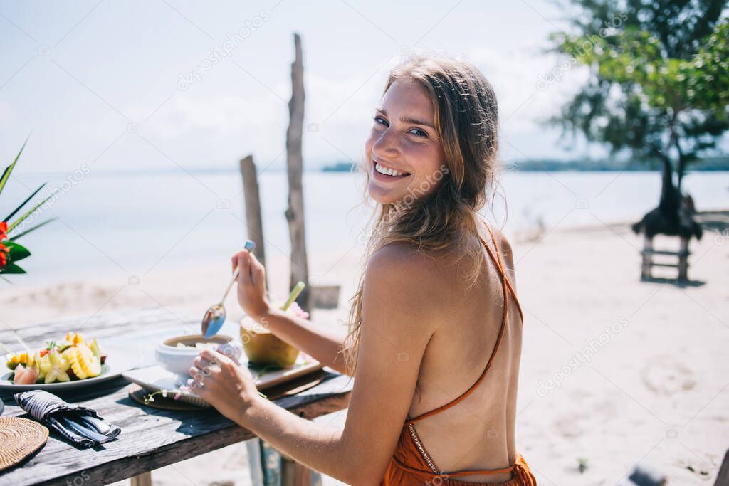 Back view of satisfied young lady in light wear sitting at wooden table and delighting in healthy food while looking at camera at empty seaside