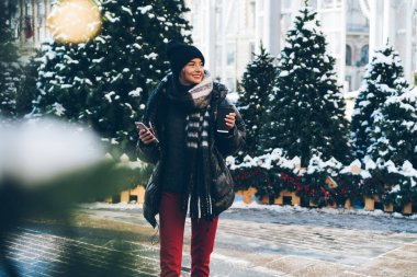 Kış kıyafetleri içinde şık bir bayan elinde kağıt bardak ve akıllı telefonuyla Noel ağaçlarının yanında dikilip sokağa bakıyor.