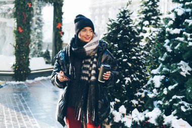 Kağıt bardak ve telefonuyla son moda kışlık kıyafetleriyle gülümseyen genç bayan Noel ağaçlarının yanında dikiliyor ve karlı caddeye bakıyor.