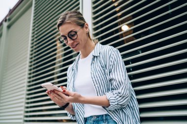 Yukardaki neşeli bayandan, günlük kıyafetlerle sosyal medyaya akıllı telefondan bakıyor ve pencereye yaslanırken gülümsüyor.