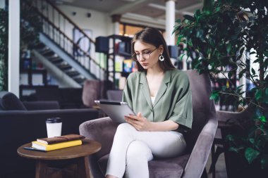 Gözlüklü, rahat koltukta oturan ve modern kütüphanede çalışmak için tablet kullanan odaklanmış genç kız öğrenci.