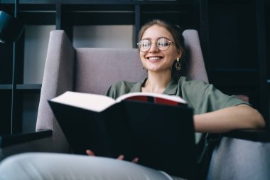 Gözlüklü mutlu bayan öğrenci kitaplıkların yanındaki modern kütüphanenin sandalyesinde otururken siyah kapaklı büyük bir kitap okuyor. 