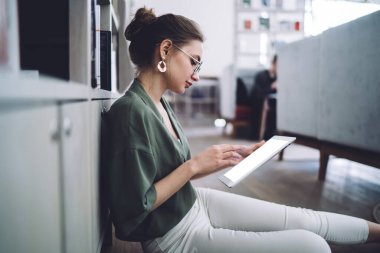 Kütüphane zemininde oturan ve tabletle çalışırken kitaplığa yaslanan gündelik giyinen genç kız öğrencinin yan görüntüsü