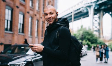 Tüylü, kulaklıklı, sırt çantalı, siyah spor kıyafetli, cep telefonunu gün ışığında kullanan ve bulanık şehir geçmişine bakan kel bir adam.