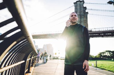 Gülümseyen atletik, traşsız, siyah spor giyim giyen genç adam şehir köprüsünün arka planında arka planda metal çitlerde akıllı telefonla iletişim kuruyor.