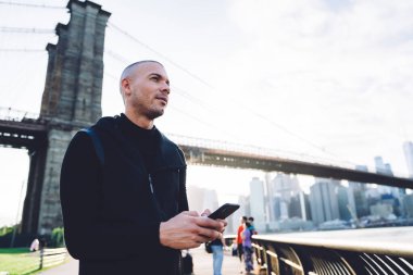 Siyah spor giyim giymiş konsantre atletik genç adam metal çitlerin oradaki barajda akıllı telefonu dinliyor ve şehir köprüsünde gün ışığına bakıyor.