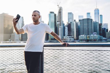 Spor kıyafetleri içinde, gökdelenlerin arka planında duran, korkuluklara tutunan ve akıllı telefondan selfie çeken genç yakışıklı bir adam.