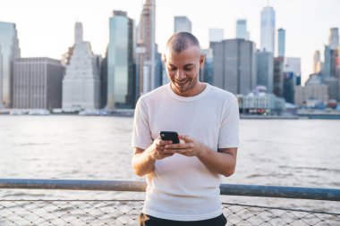 Genç, gülümseyen, kısa saçlı, beyaz tişörtlü, demir parmaklıkların yanında duran ve akıllı telefondan komik mesajlar yazan genç adam. 