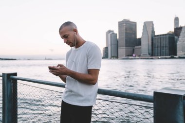 Kulaklıkla müzik dinleyen, akıllı telefon kullanan ve nehir ve şehrin arka planına zincirli çitin yanında duran genç, konsantre etnik adamın yan görüntüsü