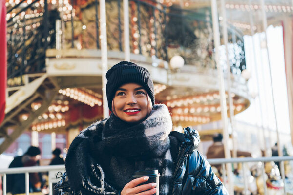 Toothy smiling young female with red lips wearing black winter hat standing by festive Christmas carousel with coffee to go looking away