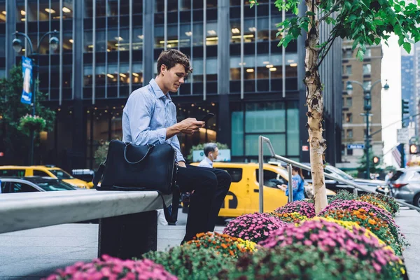 Intelligent young man in classic outfit sitting hand leaning on leather black bag surfing on cellphone beside of commercial building and colorful flowers on street with cars of New York city