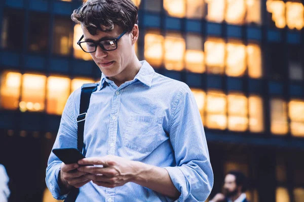 Klasik tişörtlü ve gözlüklü genç bir ofis çalışanı New York 'ta ışıklı yüksek binaya karşı telefonla mesajlaşıyor.
