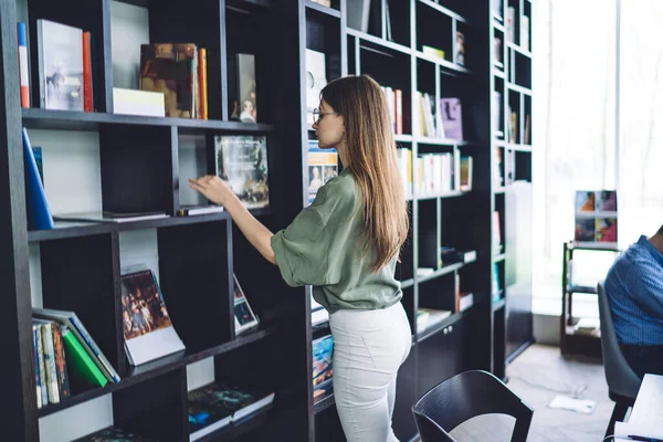 Side view of smart young woman thoughtfully checking bookshelves and choosing textbook while studying and preparing assignment in light library