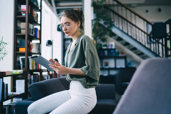 Concentrated stylish female in glasses working on tablet while sitting on armrest of couch in modern library on blurred background