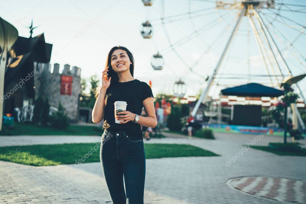 Happy Asian woman in casual wear making online international conversation via cellphone gadget connected to 4g wireless outdoors, cheerful woman with coffee to go communicated in Amusement Park