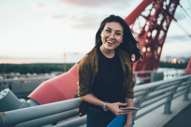 Açık havada kameraya gülümseyen, optik gözlüklü neşeli hippi kızın yarım boy portresi. Şehir merkezinde boş zamanlarında akıllı telefonlu mutlu bir kadın.