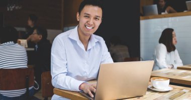 Yetişkin, neşeli, yakışıklı, zarif elbiseli Asyalı erkek. Laptop 'ta kahve içiyor ve kafeteryada kameraya bakıyor.