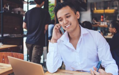 Zarif tişört giymiş, elinde kalemle telefonda konuşurken dizüstü bilgisayarın önünde oturan oldukça serbest çalışan biri.