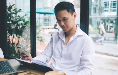 Ciddi derecede konsantre olmuş Asyalı bir adam panoramik cam pencerelerin yanındaki ahşap masada oturmuş deftere bakıyor ve telefonda daktilo yazıyor.