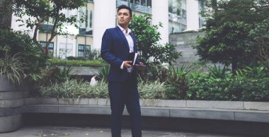 Mavi ceketli ve pantolonlu yakışıklı yetişkin işçi dışarıda yeşilliklere karşı duruyor ve elinde telefon ve planlamacı var.