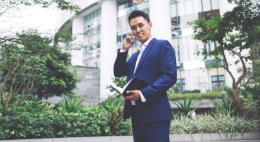 Yeşil çitlerin ve çağdaş binanın arka planında duran resmi giyinmiş, cep telefonunda kameraya bakıp gülümseyen, kendinden emin bir çalışan.