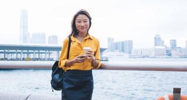 Modern kıyafetli neşeli bir etnik kadın. Sırt çantasında kahve fincanı ve akıllı bir telefonla Hong Kong rıhtımında kameraya bakarken gülümsüyor.