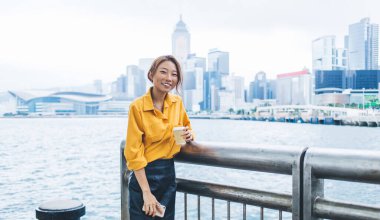 Kendine güvenen modern Asyalı kadın kahve fincanı ve akıllı telefon tutarken rüzgarlı bir günde Hong Kong rıhtımına yaslanıyor.