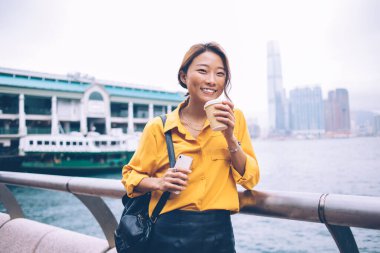 Siyah deri etekli, sırt çantalı, akıllı telefonlu, sarı tişörtlü hoş bir Asyalı kadın. Hong Kong 'da feribot iskelesinde kahve içmeye gidiyor.