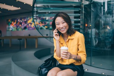 Modern koridorda oturmuş kameraya gülümserken akıllı telefondan konuşan ve gülen heyecanlı etnik kadın, Hong Kong 