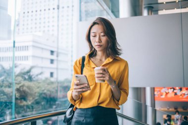 Güzel, ciddi Asyalı kadın, gündelik kıyafetleriyle modern dijital akıllı telefonuyla modern tasarımla bir bardak içkiyle geziyor ve mesajlaşıyor.
