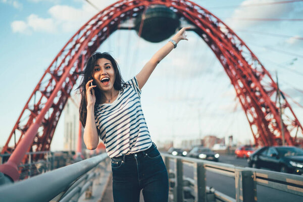 Портрет возбужденной хипстерши с поднятой рукой, потрясенной полученной новостью во время общения со смартфоном, эмоциональной женщины тысячелетия, удовлетворенной входящим международным телефонным звонком
