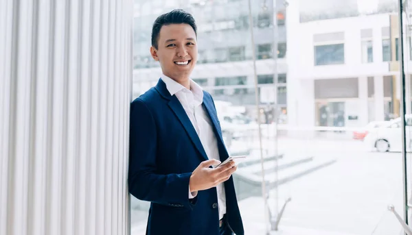 スマートフォンでスーツのテキストメッセージで幸せな笑顔アジア人男性ながら 昼光の通りにガラスオフィスビルの横にある壁にもたれて — ストック写真