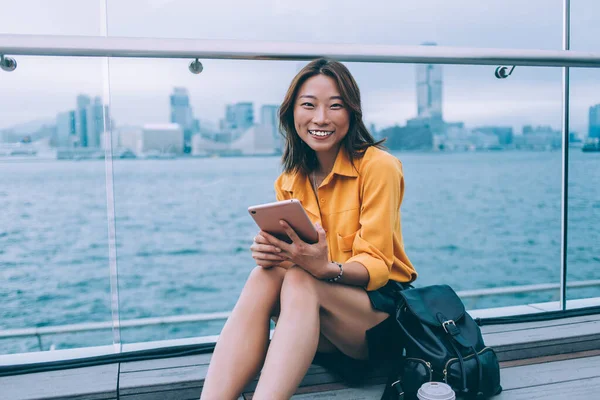 幸せな若いアジア人女性でカジュアル服で革のバックパックで香港の埠頭に座って カメラで笑顔タブレットを保持 — ストック写真