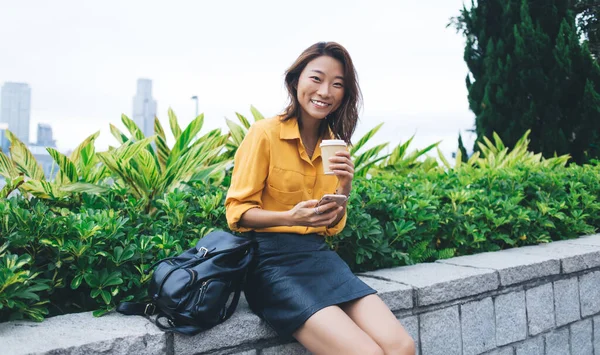 在香港街上 穿着时髦衣服 背着背包喝咖啡 拿着智能手机 坐在砖墙边的现代女性非常迷人 — 图库照片