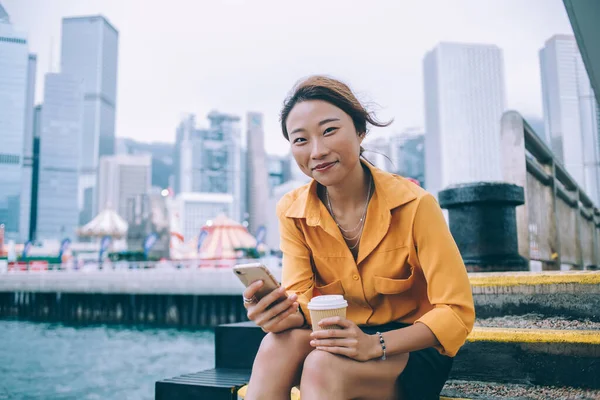 アジアの女性がコーヒーカップと携帯電話を持ちながらカメラで微笑み 香港の埠頭のコンクリートの階段に座る内容 — ストック写真