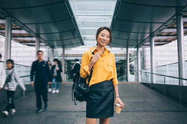コーヒーと香港の空港ターミナルに黒い革のスカートが立っている黄色のシャツを着て陽気なアジアの女性は行くとカメラを見て肩の上にバックパックを保持 — ストック写真